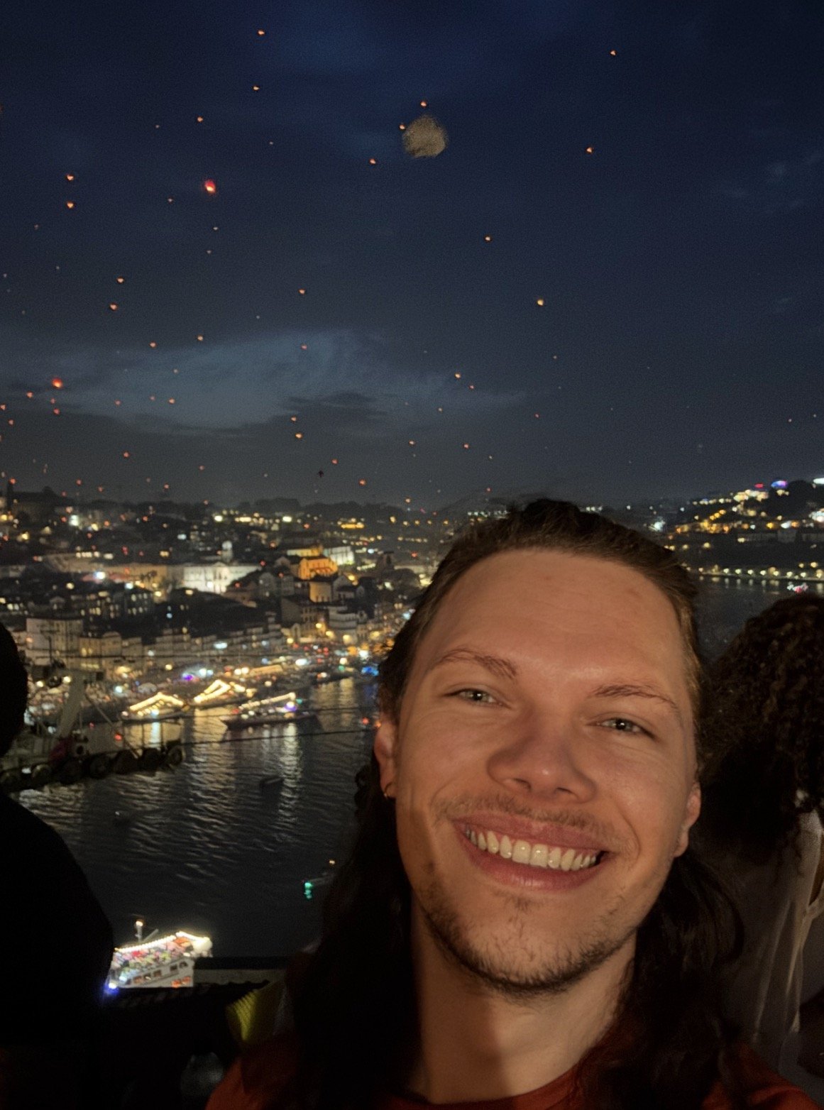 Selfie de un hombre sonriente de cabello largo, con un río, barcos y edificios iluminados al fondo. Numerosos farolillos luminosos flotan en el cielo nocturno.