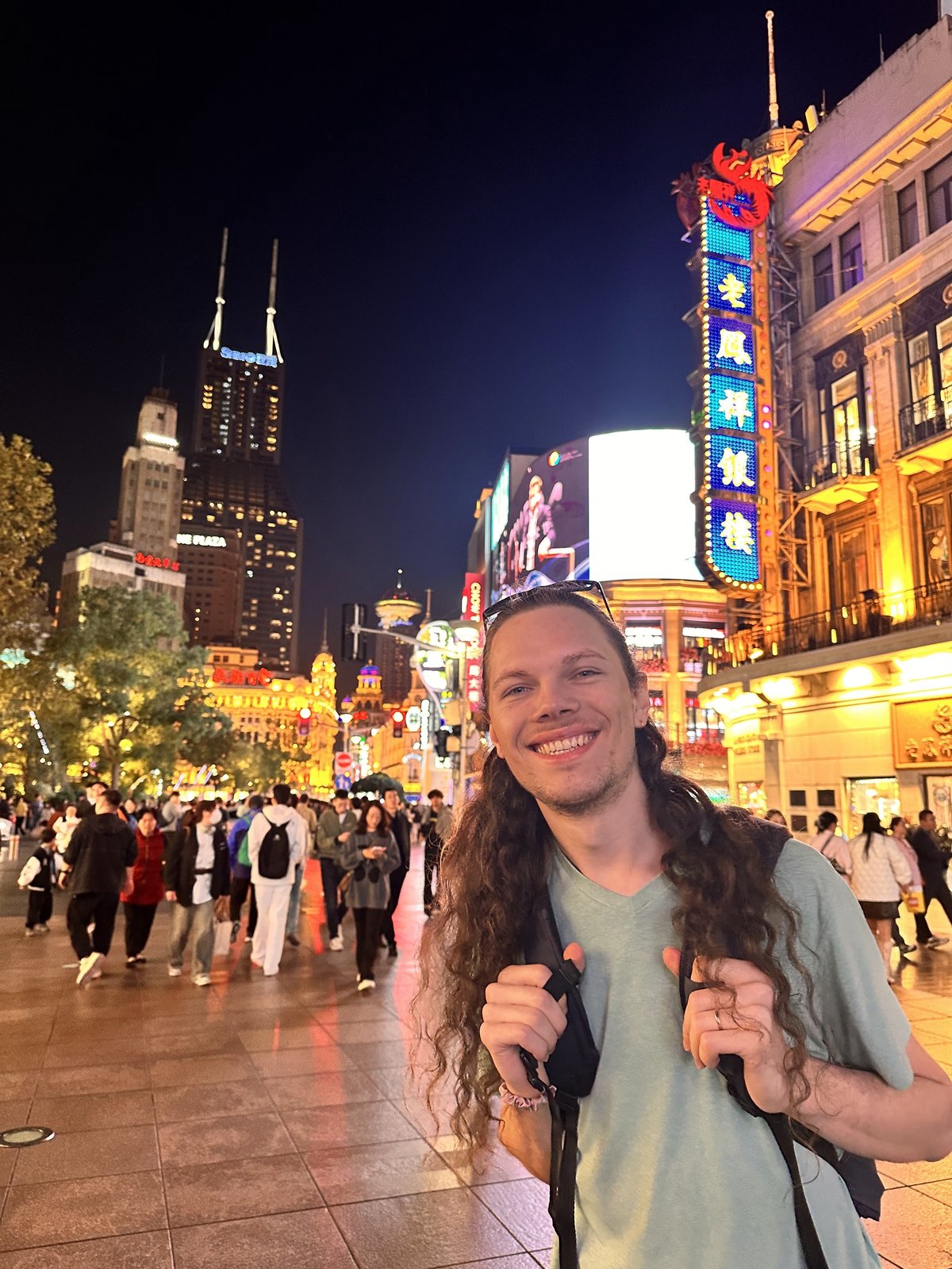 Hombre sonriente de cabello largo y camiseta verde, sujetando las correas de su mochila en una calle concurrida de noche, con edificios y letreros luminosos en chino.